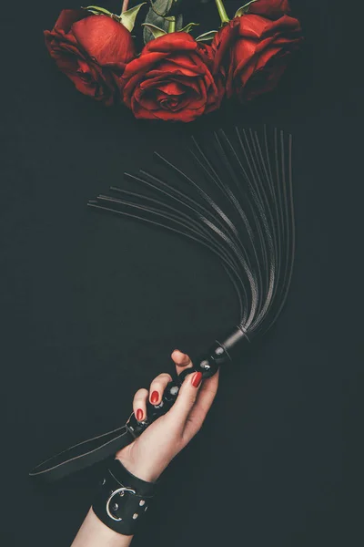 Black leather flogging whip in female hand by red roses isolated on black — Stock Photo