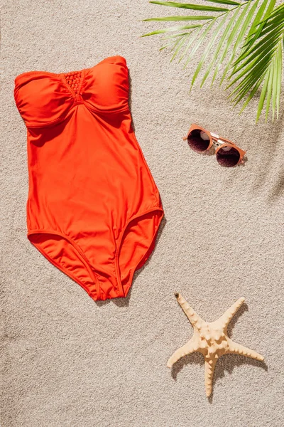 Вид сверху на стильный красный купальник и солнечные очки, лежащие на песчаном пляже — стоковое фото