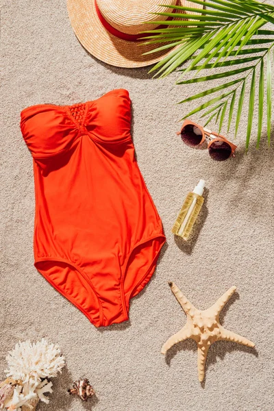 Vue de dessus du maillot de bain rouge élégant avec lotion corporelle et accessoires sur la plage de sable — Photo de stock