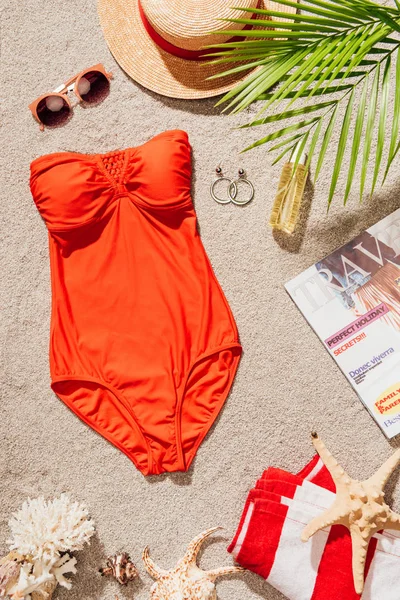 Vue de dessus du maillot de bain rouge élégant avec magazine et accessoires couchés sur la plage de sable — Photo de stock