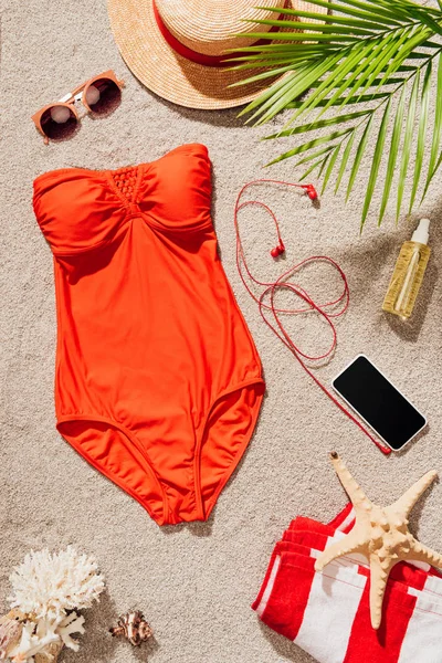 Vue de dessus du maillot de bain rouge élégant avec smartphone et accessoires couchés sur la plage de sable — Photo de stock