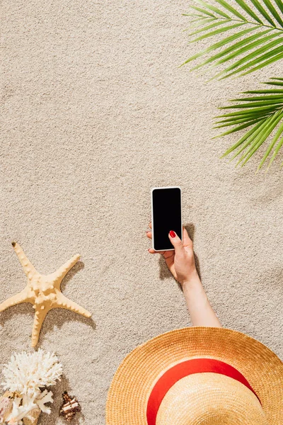 Recortado tiro de la mujer en el sombrero usando teléfono inteligente mientras se encuentra en la playa de arena - foto de stock