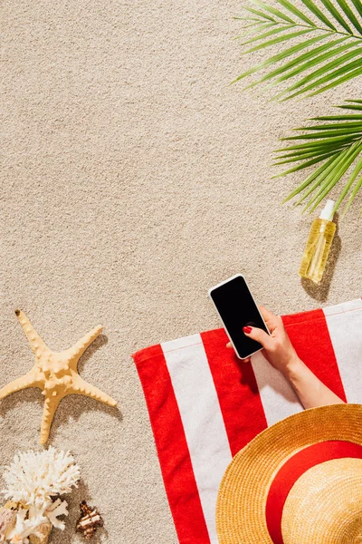 Recortado tiro de la mujer en el sombrero con el teléfono inteligente mientras se relaja en la playa de arena - foto de stock