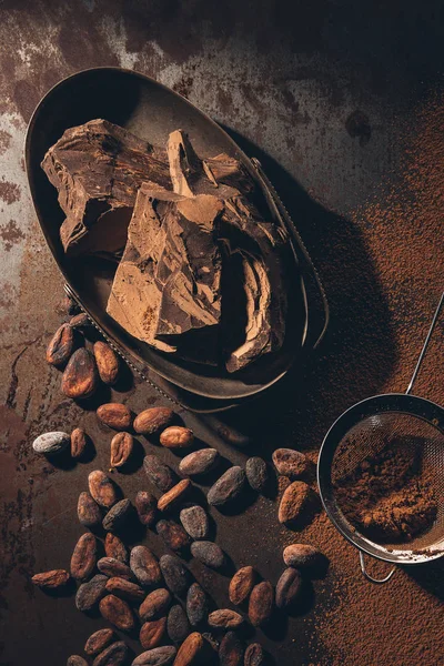 Vista superior de deliciosas peças de chocolate, grãos de cacau, pó e peneira na superfície escura — Fotografia de Stock