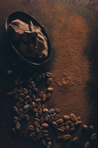 Draufsicht auf köstliche Schokoladenstücke, Kakaobohnen und Pulver auf dunkler Oberfläche — Stockfoto