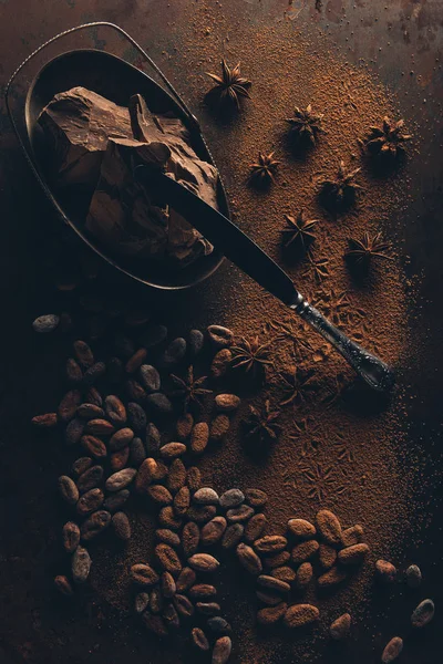 Vue de dessus des morceaux de chocolat gastronomique, couteau, fèves de cacao, poudre et anis étoilé sur la surface sombre — Photo de stock