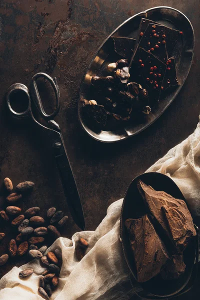 Top view of vintage scissors, cocoa beans, cloth, chocolate pieces and nuts on dark surface — Stock Photo