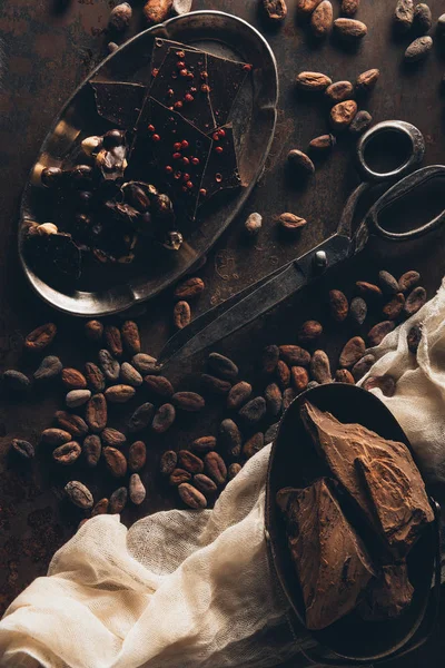 Vintage scissors, cocoa beans, cloth, chocolate pieces and nuts on dark surface — Stock Photo