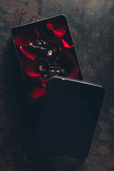 Vue du haut de la boîte avec des pétales de rose avec des morceaux de chocolat et des noisettes sur une surface sombre — Photo de stock