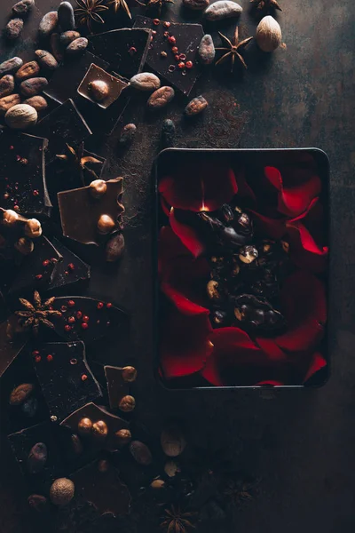 Top view of red rose petals in box with delicious chocolate pieces, nuts and cocoa beans on dark surface — Stock Photo