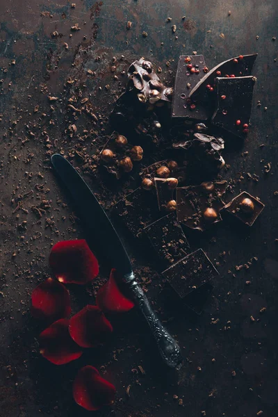 Top view of delicious chocolate pieces with nuts, vintage knife and red rose petals on dark surface — Stock Photo