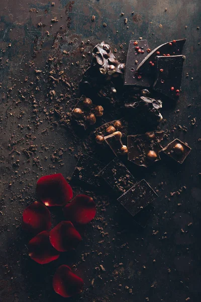 Draufsicht auf köstliche Schokoladenstücke mit Nüssen und roten Rosenblättern auf dunkler Oberfläche — Stockfoto