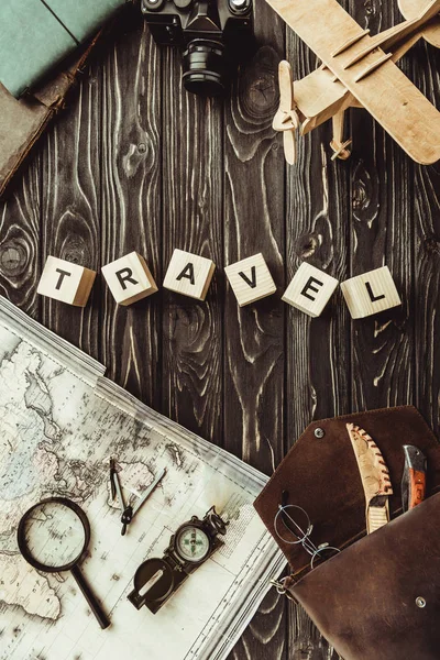 Top view of arranged wooden blocks in travel word with map, retro photo camera, bag and magnifying glass on dark wooden tabletop — Stock Photo