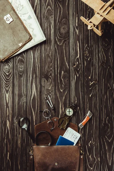 Top view of arrangement of wooden toy plane, bag with magnifying glasses, pocket knife, passport, ticket, eyeglasses and compass on dark wooden tabletop — Stock Photo