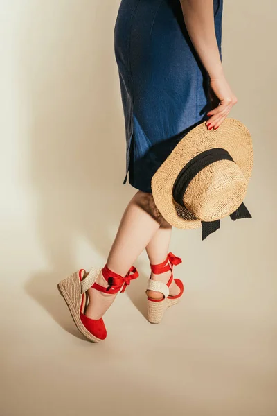 Image recadrée de femme élégante en sandales à plate-forme rouge tenant chapeau de paille sur fond beige — Photo de stock