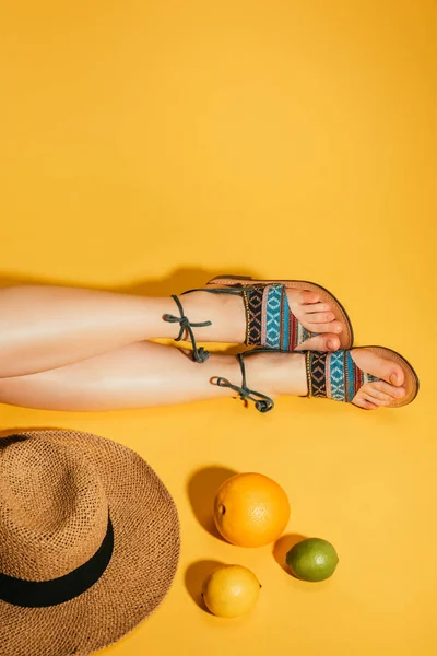 Abgeschnittene Zitrusfrüchte, Frauenfüße in stylischen Sandalen und Strohhut auf gelbem Hintergrund — Stockfoto