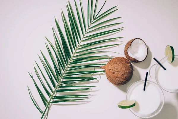 Вид зверху на окуляри з кокосовими коктейлями, лаймом та соломкою для пиття, зеленим листям долоні та екзотичними кокосами на білому — стокове фото