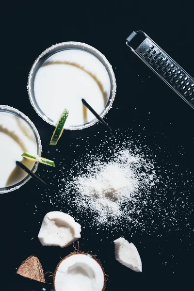 Vue du dessus des verres avec cocktails à la noix de coco et pailles à boire, râpe et copeaux de noix de coco sur noir — Photo de stock