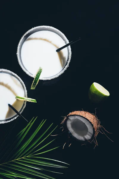 Vista dall'alto di bicchieri con cocktail di cocco e cannucce da bere, foglie di palma verde e lime su nero — Foto stock