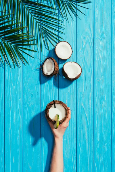 Ansicht der menschlichen Hand mit Kokoscocktail, geknackten Kokosnüssen und Palmblättern auf türkisfarbener Holzoberfläche — Stockfoto