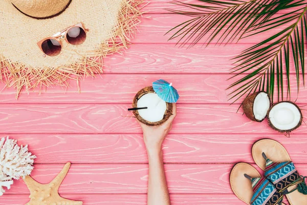 Tiro recortado de mujer sosteniendo coctel de coco rodeado de varios atributos de viaje tropicales en la superficie de madera rosa - foto de stock