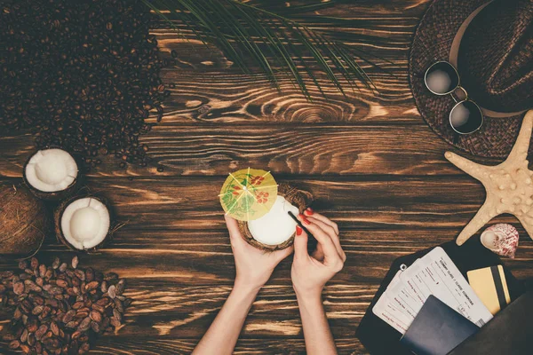 Tiro recortado de mujer con coctel de coco rodeado de varios atributos de viaje tropical en la superficie de madera - foto de stock