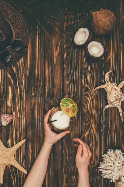 Tiro recortado de mulher com coquetel de coco e charuto na superfície de madeira cercada com vários atributos de viagem tropical — Fotografia de Stock
