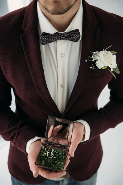 Partial view of groom in suit with buttonhole and wedding rings in box in hands — Stock Photo