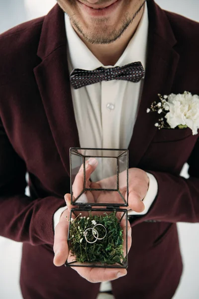 Partial view of groom in suit with buttonhole and wedding rings in box in hands — Stock Photo