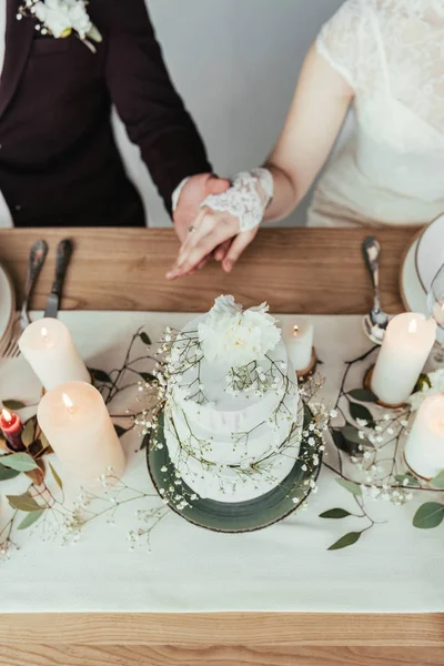 Partial view of newlyweds holding hands while sitting at served table with wedding cake, rustic wedding concept — Stock Photo