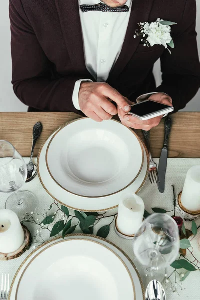 Обрезанный снимок жениха с помощью смартфона за обслуживаемым деревенским столом — стоковое фото