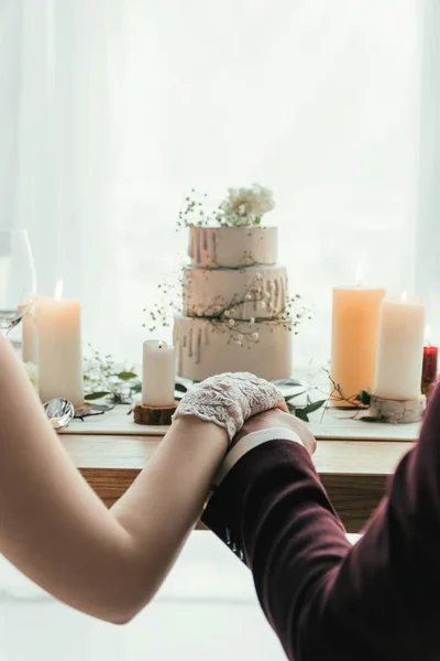 Teilansicht des Brautpaares Händchenhaltend am servierten Tisch mit Hochzeitstorte, rustikales Hochzeitskonzept — Stockfoto