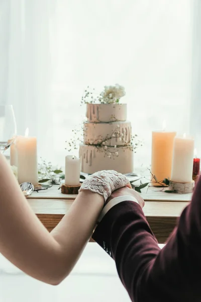 Partial view of newlyweds holding hands while sitting at served table with wedding cake, rustic wedding concept — Stock Photo