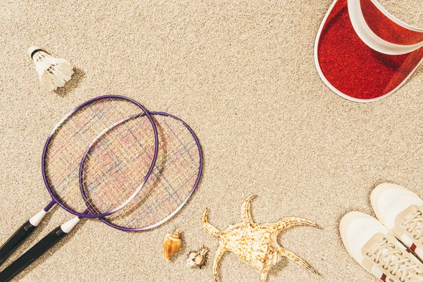 Vue de dessus des équipements, chaussures, casquettes et coquillages de badminton sur le sable — Photo de stock