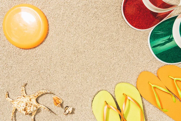Tendido plano con disco volador, tapas de colores, chanclas y conchas marinas en la arena - foto de stock