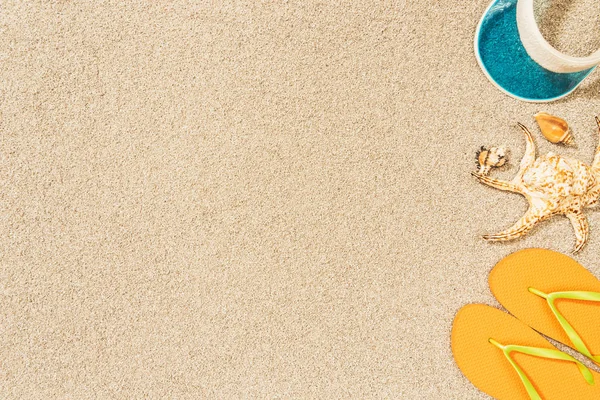 Tendido plano con disposición de conchas marinas, chanclas amarillas y gorra azul sobre arena - foto de stock