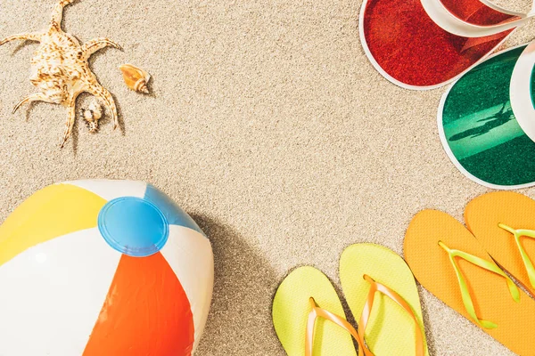 Flat lay with colorful flip flops, beach ball, seashells and caps arranged on sand — Stock Photo