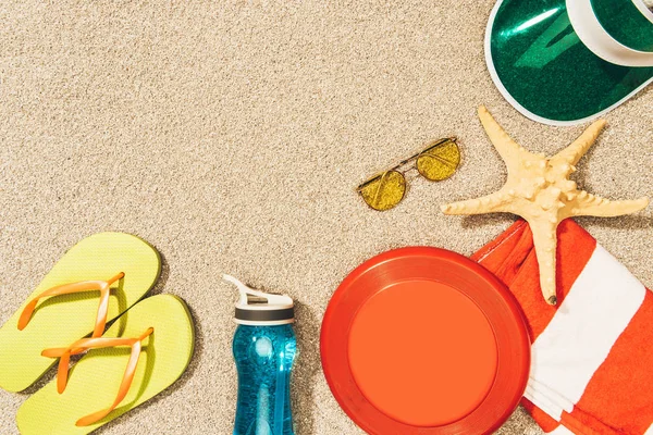 Flat lay with frisbee, cap, flip flops, sunglasses and water bottle arranged on sand — Stock Photo