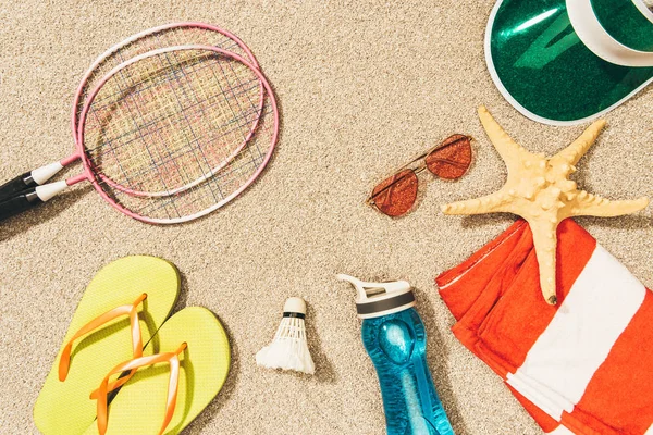 Blick von oben auf Badminton-Ausrüstung, Sonnenbrille, Flip Flops, Mütze und Handtuch auf Sand — Stockfoto