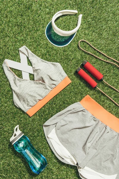 Вид сверху на спортивную одежду, бутылку воды, скакалку и шапку на зеленой траве — стоковое фото