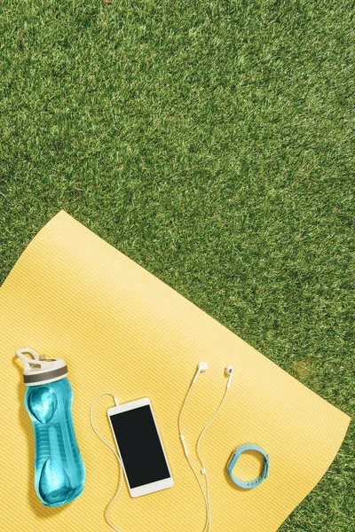 Cama plana con botella de agua, reloj, teléfono inteligente y auriculares en la estera amarilla en el césped verde - foto de stock