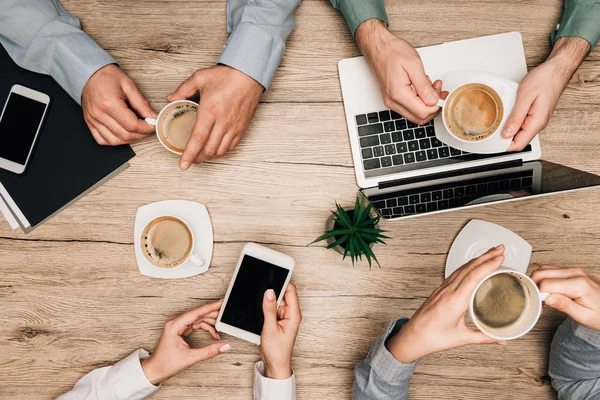 Top view of business partners drinking coffee by gadgets on table, cropped view — Stock Photo