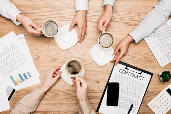 Top view of businesswomen and businessman drinking coffee by contract and documents on table — Stock Photo