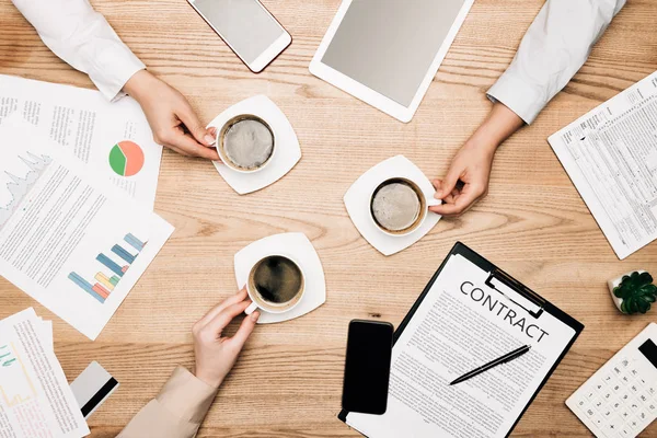 Top view of business partners drinking coffee by paperwork and contract on wooden table — Stock Photo