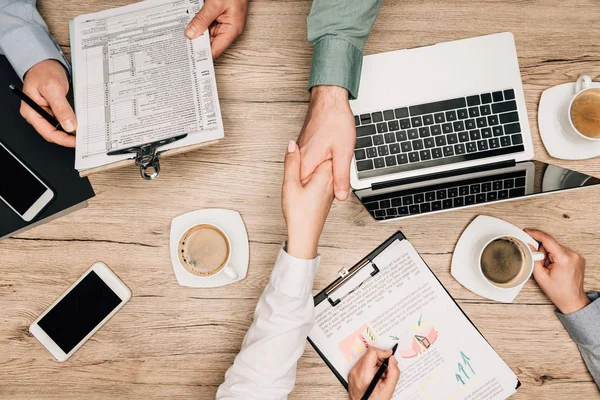 Vista superior de la gente de negocios estrechando la mano por documentos, ordenador portátil y café en la mesa - foto de stock