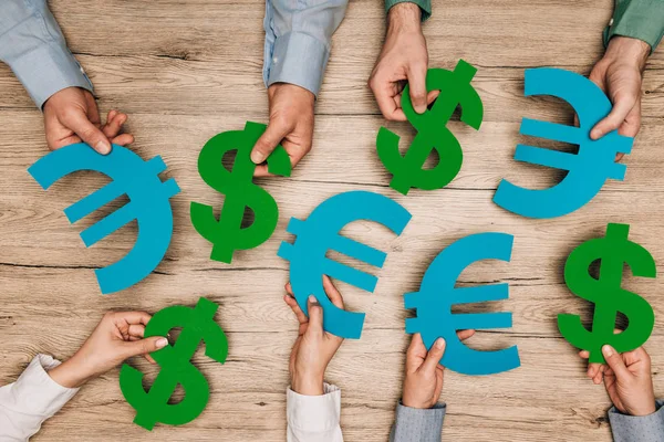 Top view of business people holding dollar and euro signs on wooden table, cropped view — Stock Photo