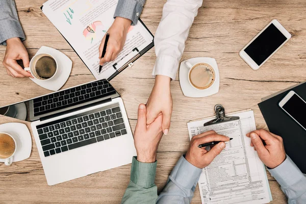 Top view of business people handshaking by gadgets, documents and coffee on table — Stock Photo