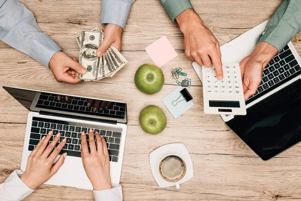 Вид сверху деловых людей, работающих с ноутбуком, калькулятором и считающих долларовые банкноты за столом — стоковое фото