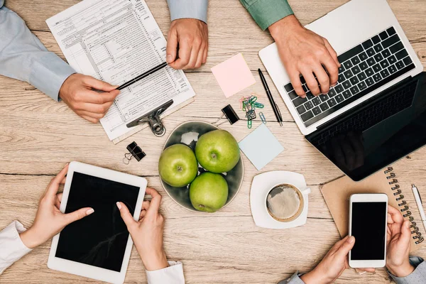 Vista superior de socios comerciales con dispositivos digitales, documentos y papelería sobre la mesa, vista recortada - foto de stock