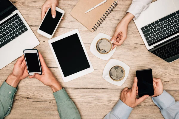 Top view of business partners using gadgets beside coffee on table, cropped view — Stock Photo